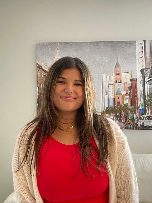 A picture of Elise Mendelsohn, who wears a red shirt, beige sweater, and poses inside.