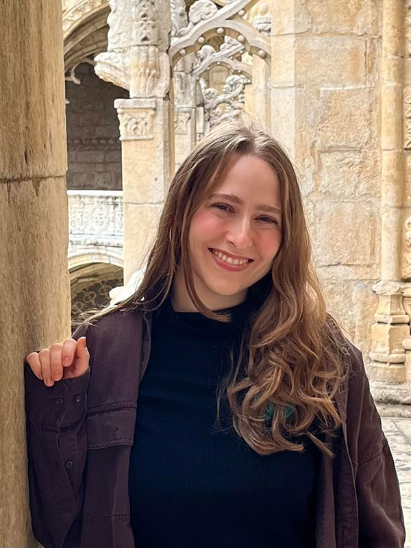 A headshot of Natanya Katz, who wears a black shirt, brown jacket, and poses outdoors.